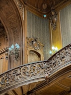Intricately carved wooden paneling and railing adorn an interior space, featuring ornate designs and patterns. Soft, ambient lighting is provided by elegant lamps with pale blue light bulbs, complementing the rich textures of the woodwork. The walls are covered with subtle damask wallpaper, adding to the opulent atmosphere.