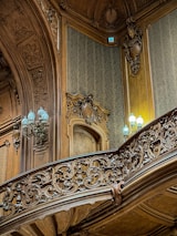 Intricately carved wooden paneling and railing adorn an interior space, featuring ornate designs and patterns. Soft, ambient lighting is provided by elegant lamps with pale blue light bulbs, complementing the rich textures of the woodwork. The walls are covered with subtle damask wallpaper, adding to the opulent atmosphere.