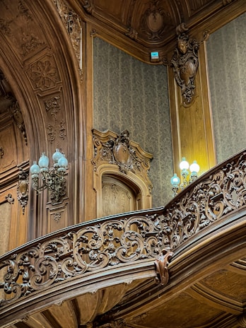 Intricately carved wooden paneling and railing adorn an interior space, featuring ornate designs and patterns. Soft, ambient lighting is provided by elegant lamps with pale blue light bulbs, complementing the rich textures of the woodwork. The walls are covered with subtle damask wallpaper, adding to the opulent atmosphere.