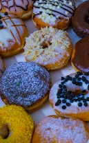 A vibrant assortment of glazed donuts with various toppings, including chocolate drizzle, white chocolate chips, powdered sugar, and colorful sprinkles. The donuts are arranged closely together, showcasing a mix of textures and flavors.