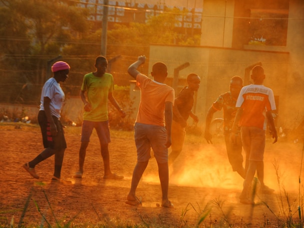 A group of young people are playing football on a dusty field during sunset. The warm, golden light casts dramatic shadows and highlights the dust stirred up by their movements. The scene suggests energy and camaraderie.
