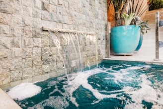 Water cascades from a narrow spout along a stone wall into a bubbling pool below. A large turquoise planter with lush green plants is positioned nearby, offering a tranquil and refreshing atmosphere.