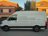 A large white delivery van is parked on a street in front of two-story houses made of red brick with beige trim. The houses have tiled roofs, and one has a tall evergreen tree adjacent to it. The lighting suggests a late afternoon or early evening setting.