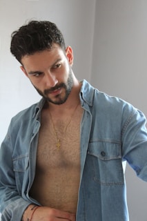 A man with short dark hair and a beard wears an unbuttoned denim shirt, revealing a bare chest and a gold cross necklace. He has a contemplative expression and stands against a plain background.