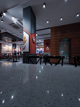 A modern fast-food restaurant interior features sleek lighting fixtures and wooden accents. The space includes tables and chairs arranged neatly. Wall decorations include a large logo and branded signage, creating a branded atmosphere.