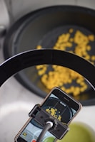 An overhead view shows a smartphone mounted on a stand recording yellow corn kernels in a dark container. The phone's screen displays a live video feed of the kernels.