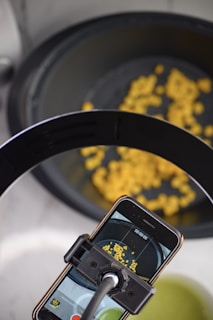 An overhead view shows a smartphone mounted on a stand recording yellow corn kernels in a dark container. The phone's screen displays a live video feed of the kernels.