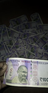 A close-up of a person holding a 100 Indian Rupees banknote prominently displays the note's design, featuring Mahatma Gandhi's portrait. In the background, multiple 100 Rupees banknotes are laid out, suggesting a substantial quantity of money.