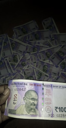 A close-up of a person holding a 100 Indian Rupees banknote prominently displays the note's design, featuring Mahatma Gandhi's portrait. In the background, multiple 100 Rupees banknotes are laid out, suggesting a substantial quantity of money.