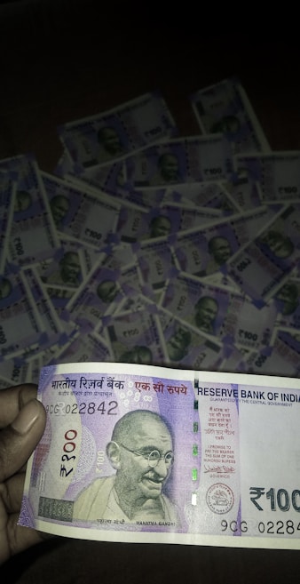 A close-up of a person holding a 100 Indian Rupees banknote prominently displays the note's design, featuring Mahatma Gandhi's portrait. In the background, multiple 100 Rupees banknotes are laid out, suggesting a substantial quantity of money.