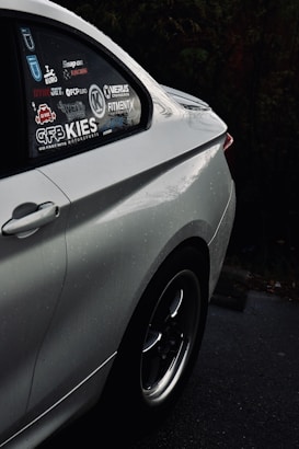 The image displays the rear side of a silver car parked on a wet surface. The car's window is adorned with multiple decals and logos related to cars and motorsports, suggesting an interest in automotive tuning or modification. Raindrops are visible on the car's surface, indicating recent rainfall. The background is dark, possibly due to the overcast weather or the time of day.
