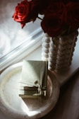 A marble tray holds a neatly wrapped Gucci gift box, tied with a ribbon. Next to it, a vase crafted from spherical beads is filled with vibrant red roses. Soft sunlight filters through a window, casting gentle shadows across the scene.