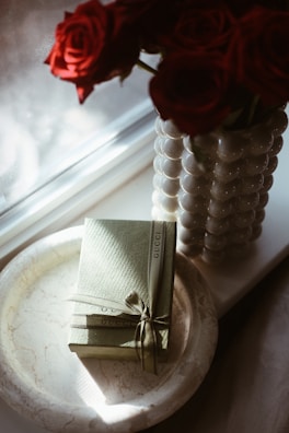 A marble tray holds a neatly wrapped Gucci gift box, tied with a ribbon. Next to it, a vase crafted from spherical beads is filled with vibrant red roses. Soft sunlight filters through a window, casting gentle shadows across the scene.