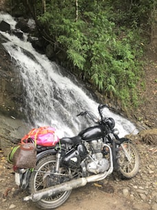 A black motorcycle is parked in front of a cascading waterfall surrounded by lush greenery. The motorcycle has a brown leather bag and a vibrant pink and orange backpack strapped to it. The terrain is rocky and earthy, adding to the natural setting.