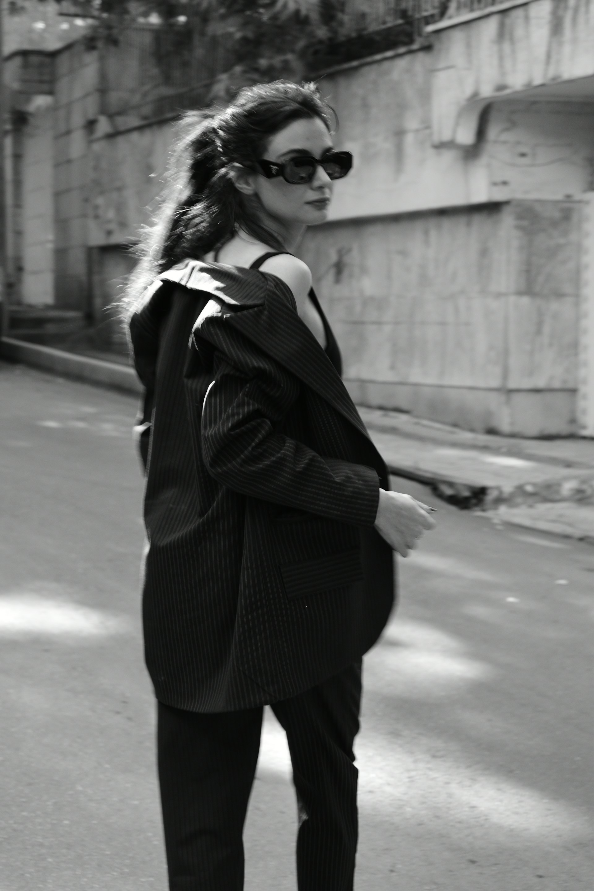 A woman with long hair wearing sunglasses and an oversized pinstripe suit jacket walks down an empty urban street. The photograph is in black and white, giving it a timeless and classic feel.