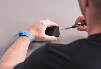 A person is repairing a smartphone with a damaged screen. They hold a screwdriver while wearing an anti-static wristband.
