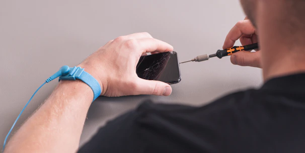 A person is repairing a smartphone with a damaged screen. They hold a screwdriver while wearing an anti-static wristband.