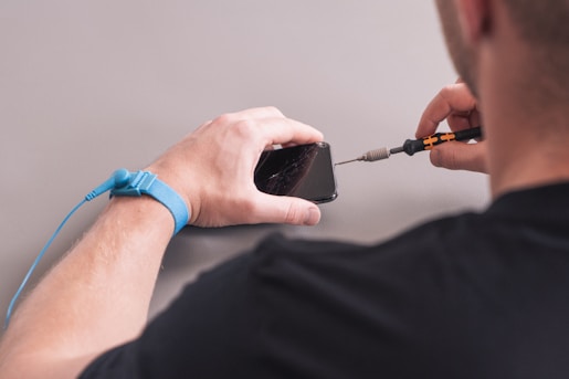 A person is repairing a smartphone with a damaged screen. They hold a screwdriver while wearing an anti-static wristband.