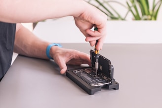 A person is working on an open smartphone, using a screwdriver to repair or modify the device. The phone is positioned on a mat, with the screen lifted, exposing the internal components. A blue wristband watch is visible on the person's wrist, and a part of a plant is blurred in the background.