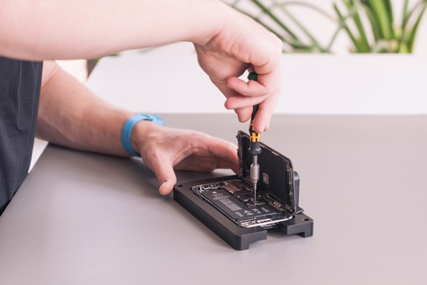 A person is working on an open smartphone, using a screwdriver to repair or modify the device. The phone is positioned on a mat, with the screen lifted, exposing the internal components. A blue wristband watch is visible on the person's wrist, and a part of a plant is blurred in the background.