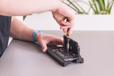 A person is working on an open smartphone, using a screwdriver to repair or modify the device. The phone is positioned on a mat, with the screen lifted, exposing the internal components. A blue wristband watch is visible on the person's wrist, and a part of a plant is blurred in the background.