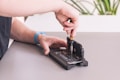 A person is working on an open smartphone, using a screwdriver to repair or modify the device. The phone is positioned on a mat, with the screen lifted, exposing the internal components. A blue wristband watch is visible on the person's wrist, and a part of a plant is blurred in the background.