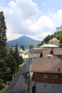 A picturesque view of a small town nestled in a valley with a mountainous backdrop. There are multiple buildings with rustic roofs surrounded by lush greenery. A castle is perched atop a hill on the right, adding a historical element to the scene. The sky is partly cloudy, creating a serene atmosphere.