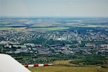An aerial view showcasing an expansive landscape with a combination of urban and rural elements. Industrial facilities and residential buildings are visible alongside fields and open land. Smoke can be seen rising from one of the industrial chimneys. The mixture of natural greenery and man-made structures creates a diverse setting.