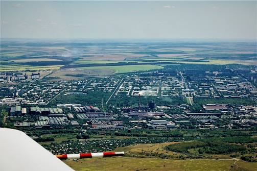 An aerial view showcasing an expansive landscape with a combination of urban and rural elements. Industrial facilities and residential buildings are visible alongside fields and open land. Smoke can be seen rising from one of the industrial chimneys. The mixture of natural greenery and man-made structures creates a diverse setting.