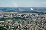 Aerial view of a city with dense green areas surrounding numerous residential and commercial buildings. In the background, expansive agricultural lands with clear demarcation are visible, showcasing a mix of green and brown fields.