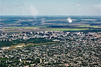 Aerial view of a city with dense green areas surrounding numerous residential and commercial buildings. In the background, expansive agricultural lands with clear demarcation are visible, showcasing a mix of green and brown fields.
