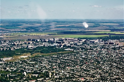 Aerial view of a city with dense green areas surrounding numerous residential and commercial buildings. In the background, expansive agricultural lands with clear demarcation are visible, showcasing a mix of green and brown fields.