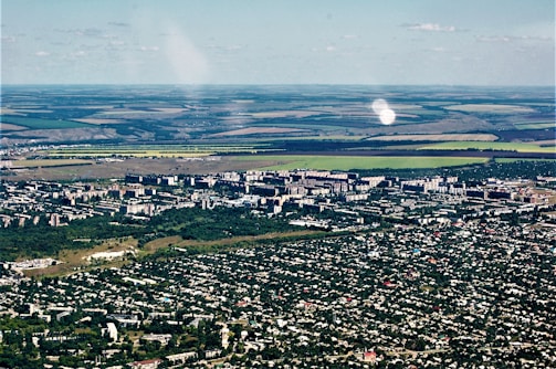 Aerial view of a city with dense green areas surrounding numerous residential and commercial buildings. In the background, expansive agricultural lands with clear demarcation are visible, showcasing a mix of green and brown fields.