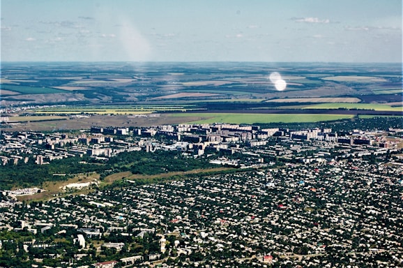 Aerial view of a city with dense green areas surrounding numerous residential and commercial buildings. In the background, expansive agricultural lands with clear demarcation are visible, showcasing a mix of green and brown fields.