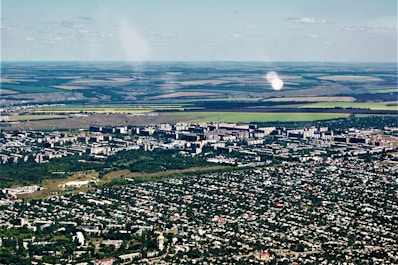 Aerial view of a city with dense green areas surrounding numerous residential and commercial buildings. In the background, expansive agricultural lands with clear demarcation are visible, showcasing a mix of green and brown fields.
