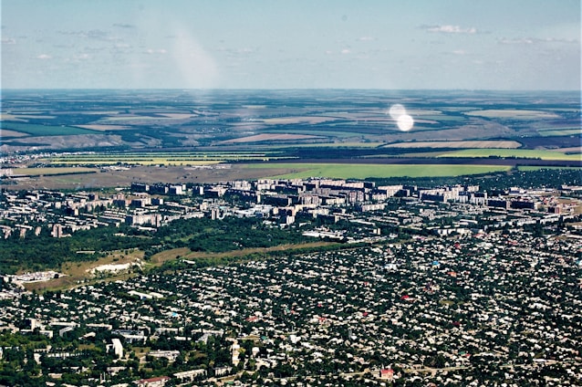 Aerial view of a city with dense green areas surrounding numerous residential and commercial buildings. In the background, expansive agricultural lands with clear demarcation are visible, showcasing a mix of green and brown fields.
