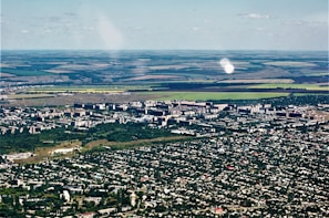 Aerial view of a city with dense green areas surrounding numerous residential and commercial buildings. In the background, expansive agricultural lands with clear demarcation are visible, showcasing a mix of green and brown fields.