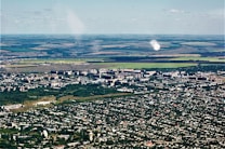 Aerial view of a city with dense green areas surrounding numerous residential and commercial buildings. In the background, expansive agricultural lands with clear demarcation are visible, showcasing a mix of green and brown fields.