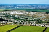 An aerial view of a rural landscape featuring large expanses of agricultural fields, a collection of houses forming a small town or village, and a backdrop of rolling hills. The fields are predominantly green with some areas appearing brown, possibly indicating different crop stages or types. A distinct section of white, possibly chalk or rock, is visible on the hills, adding contrast to the scene.