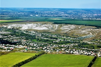An aerial view of a rural landscape featuring large expanses of agricultural fields, a collection of houses forming a small town or village, and a backdrop of rolling hills. The fields are predominantly green with some areas appearing brown, possibly indicating different crop stages or types. A distinct section of white, possibly chalk or rock, is visible on the hills, adding contrast to the scene.
