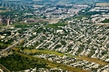 An aerial view of a sprawling urban and industrial area surrounded by extensive greenery. Residential neighborhoods with numerous houses are prominent in the foreground, while industrial facilities and factories are visible in the background. Lush trees and vegetation are interspersed throughout the cityscape, creating a stark contrast between urban development and nature.