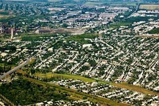 An aerial view of a sprawling urban and industrial area surrounded by extensive greenery. Residential neighborhoods with numerous houses are prominent in the foreground, while industrial facilities and factories are visible in the background. Lush trees and vegetation are interspersed throughout the cityscape, creating a stark contrast between urban development and nature.