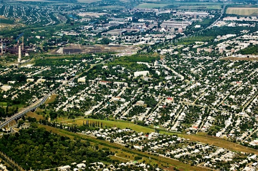 An aerial view of a sprawling urban and industrial area surrounded by extensive greenery. Residential neighborhoods with numerous houses are prominent in the foreground, while industrial facilities and factories are visible in the background. Lush trees and vegetation are interspersed throughout the cityscape, creating a stark contrast between urban development and nature.