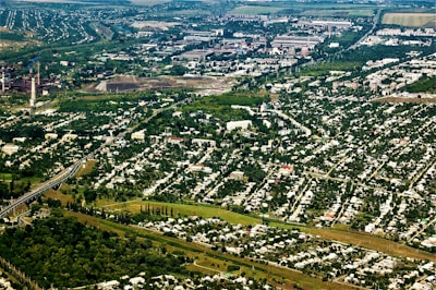 An aerial view of a sprawling urban and industrial area surrounded by extensive greenery. Residential neighborhoods with numerous houses are prominent in the foreground, while industrial facilities and factories are visible in the background. Lush trees and vegetation are interspersed throughout the cityscape, creating a stark contrast between urban development and nature.