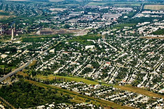 An aerial view of a sprawling urban and industrial area surrounded by extensive greenery. Residential neighborhoods with numerous houses are prominent in the foreground, while industrial facilities and factories are visible in the background. Lush trees and vegetation are interspersed throughout the cityscape, creating a stark contrast between urban development and nature.