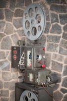 A vintage film projector is positioned against a rough stone wall. The device features large metal spools and intricate mechanical components with dials and buttons, suggesting a classic design from an earlier era of cinema.