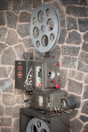 A vintage film projector is positioned against a rough stone wall. The device features large metal spools and intricate mechanical components with dials and buttons, suggesting a classic design from an earlier era of cinema.