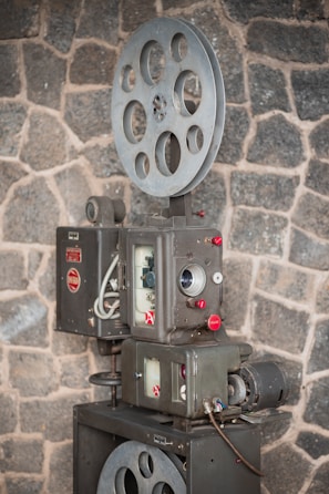 A vintage film projector is positioned against a rough stone wall. The device features large metal spools and intricate mechanical components with dials and buttons, suggesting a classic design from an earlier era of cinema.