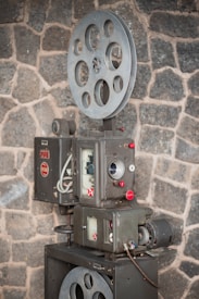 A vintage film projector is positioned against a rough stone wall. The device features large metal spools and intricate mechanical components with dials and buttons, suggesting a classic design from an earlier era of cinema.