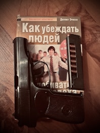 A book with a cover featuring a smiling person is placed on a wooden surface. A handgun is resting on the book, partially covering the title and image.